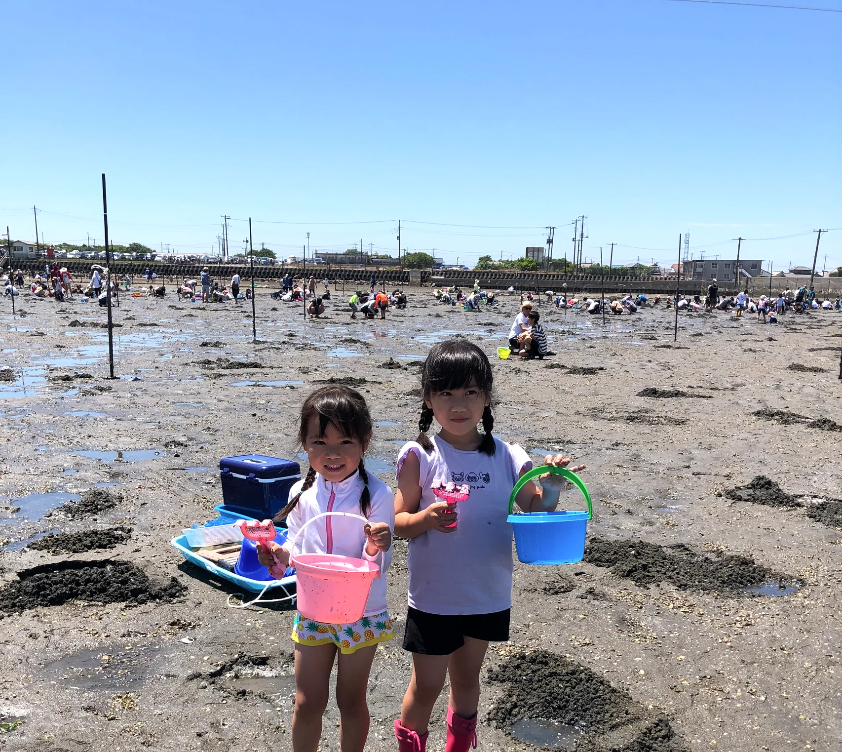 青空の牛込海岸で、バケツと熊手を持って笑顔を見せる女の子2人
